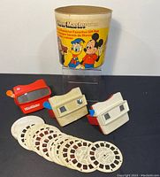 Wide shot of three vintage View-Master viewers and multiple reels with a View-Master packaging tube in the background.