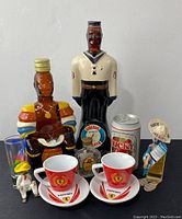 Front view of vintage barware bottles including sailor shaped bottle, bird shaped bottle, miniatures, Ferrari cups and saucers, and decorative figurines arranged on black surface.