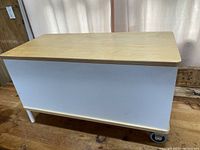 Front view of white storage bench with natural wood top and visible wheel.