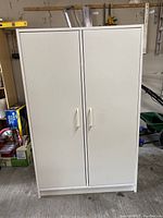 Closed white cabinet with double doors and handles, standing in a garage with tools and boxes in background.