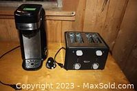Coffee maker and black four-slot toaster next to each other on wooden surface, showing front and side views.