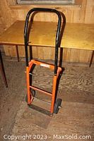 Full view of the manual moving dolly showing black handle and orange frame beside a wooden table