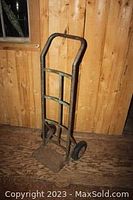 Front view of an old metal hand dolly leaning against a wooden wall on a wooden floor.