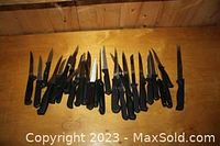 View showing a large assortment of steak knives and kitchen cutting tools laid out on a wooden surface, black handles, various blade sizes and styles.