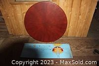 Photo showing round wooden table top with reddish-brown finish and rectangular plywood sign with painted illustration and text on blue background.