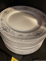 Stack of 12 white dinner plates with floral blue and gray border design and gold rim.