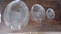 Three clear glass platters shaped like fish arranged by size on wood surface