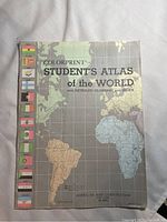 Front cover of the atlas showing world map and multiple country flags along the left edge. Title 'Colorprint Student's Atlas of the World' visible with publisher details dated 1953.