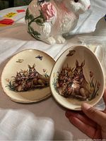 Close-up of two ceramic egg-shaped plates with bunny illustrations and floral motifs; one plate shows worn rim