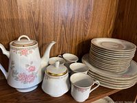 Full view of set displayed on wooden shelf showing teapot, sugar bowl, cups, and stacked plates with floral pink rose and gold trim design.
