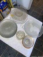 Photo showing multiple clear glass dishes and platters including textured fruit platter, patterned larger platters, saucers, and a glass vase arranged on a white cloth.