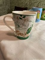 Photo showing a white ceramic coffee mug with green and brown floral design, with other character design mugs visible in the background.