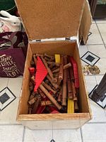 Open cardboard box showing assorted Lincoln Logs wooden pieces including natural wood logs and red and yellow roof pieces.