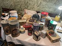 Photo showing a collection of 23 vintage decorative tins of various shapes and sizes arranged on a surface against a textured background, including winter themes, food brand tins, animals, and holiday designs.