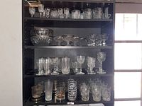 Shelf containing a wide variety of glassware including wine glasses, dessert bowls, tumblers, and goblets in clear and tinted glass with etched and textured patterns. Shelf is black, window to the right for natural light.