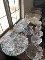Various clear crystal serving bowls, platters, covered dishes, and small decorative pieces arranged on a table near a window.