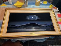Framed painting showing a dark moonlit seascape with waves and silhouetted landmass under a cloudy sky, displayed on a table with other items around.