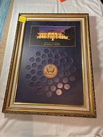 Framed display for U.S. Presidential Dollar coins showing 39 empty slots, central presidential seal emblem, and a photo of the White House at night above.