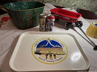 Collection of items displayed including green metal decorative bucket, two vintage soda cans, small red Radio Flyer toy cart, and white Marine Corps tray.