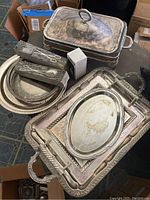 Photo of multiple silver plated trays and covered serving dish stacked together, some with ornate handles and edges