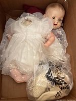 Vintage baby doll lying in a box with head partially detached. Doll wears white embroidered dress and has rosy cheeks and painted lips.