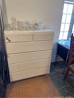 Photo showing white wooden dresser with six drawers, paint is chipped and peeling in spots indicating heavy use, with no hardware on drawers visible, placed on a wood floor near a window.