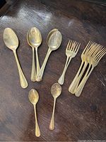 Photo shows a set of nine vintage flatware pieces with a gold-tone finish, including four forks, three large spoons, and two small spoons placed on a wooden surface.