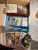 Photo showing Nintendo Wii console packaging inserts, two Wii Remote controllers with wrist straps, cables, and six Wii game cases including Wii Sports and others.