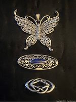 Photo showing three silver jewelry pieces: butterfly pendant with rhinestones, filigree blue stone pin, and woven silver pin.