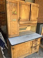 Front view of the full antique Hoosier cabinet showing wood doors, enamel work surface, drawer, and painted leaf decoration.