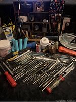 Photo showing drill bits, red handle screwdrivers, blue plastic cones, tapes, scissors, hammer, wire spool, brass fittings, and various small tools on wooden surface with shelving in background.
