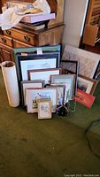 Collection of multiple framed artworks and prints, stacked and leaning against a wooden chest and wall, various frame types and sizes visible.