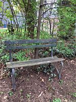Front and side views of weathered wooden outdoor bench with green metal legs and frame placed in backyard area with foliage background.