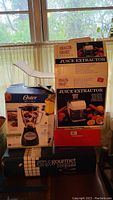 Photo showing boxed Oster Blender, Health Smart Juice Extractor, Estia frying pan box, and red box likely Chefmate sandwich maker.