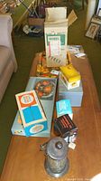Overview photo of the wooden table displaying several vintage household items including a boxed Pyrex coffee server, a vintage candle holder, metal boxes, and packaged tumblers
