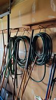 Three garden hoses coiled and hanging on wall hooks, clearly showing two round hoses and one flat soaker hose.