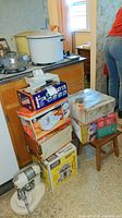 Stack of boxed kitchen appliances and slicers on stool and floor, with vintage mixers and large enamel pots on stove