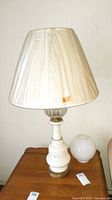 Cream ceramic and metal lamp with floral detail and pleated fabric shade on wood surface with round white glass globe in background