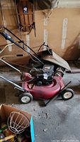 Two lawn mowers in a garage setting; Murray mower red with beige grass bag, smaller Black & Decker mower behind it.