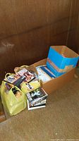 Lot with multiple yellow paper bags and a cardboard box containing vintage VHS tapes, DVDs, and magazines on floor against wooden wall.