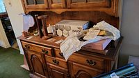 Photo showing wooden sideboard with various items on top including wine goblets boxes, wrapped items likely platters and trays, ashtrays and dishware under packing paper.