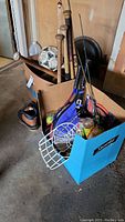 Two cardboard boxes containing various sports equipment including baseball bats, balls, mitts, a soccer ball, tennis racquets with balls, fishing poles, and men's boots.