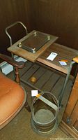 Tea cart with metal handles and wooden slats on wheels, ornate metal bucket on floor, and a wood framed mirrored lazy Susan on top of cart