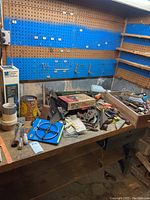 Workbench covered with assorted hand tools, tapes, instruction manuals, hardware boxes and other small tools on wooden surface with pegboard and shelves in background.