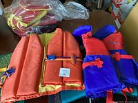 Six life jackets arranged on a table showing four orange adult jackets with blue and yellow straps and two purple and red youth jackets with red straps
