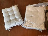 Photo shows one tufted rectangular chair seat cushion in light neutral fabric with four corner ties, placed beside a sealed package containing remaining cushions in the set.