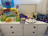 Photo showing VTech Smart Shots Sports Centre, PJ Masks electronic game, musical toy, duck pull toy and small assorted toys on a white dresser under a window