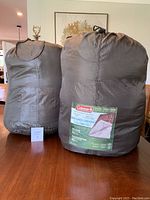 Two dark olive green Coleman sleeping bags in compression sacks, one with visible label showing brand and size.