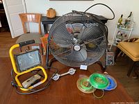 Photo of Honeywell commercial fan, yellow portable shop lamp with cord, manual crank lamp, and three spools of grass trimmer cord in tan, green, and blue colors placed on a wooden table.