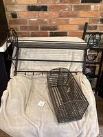 Black metal wall rack with hooks and grid shelf, placed on beige blanket against brick background.